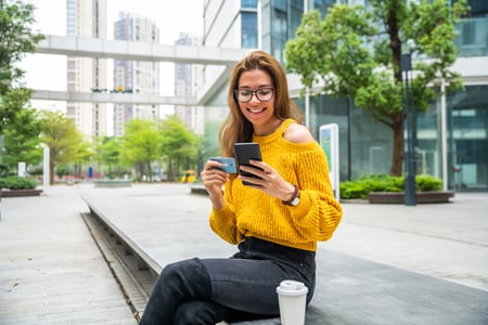 mulher numa praça usando o celular e sorrindo